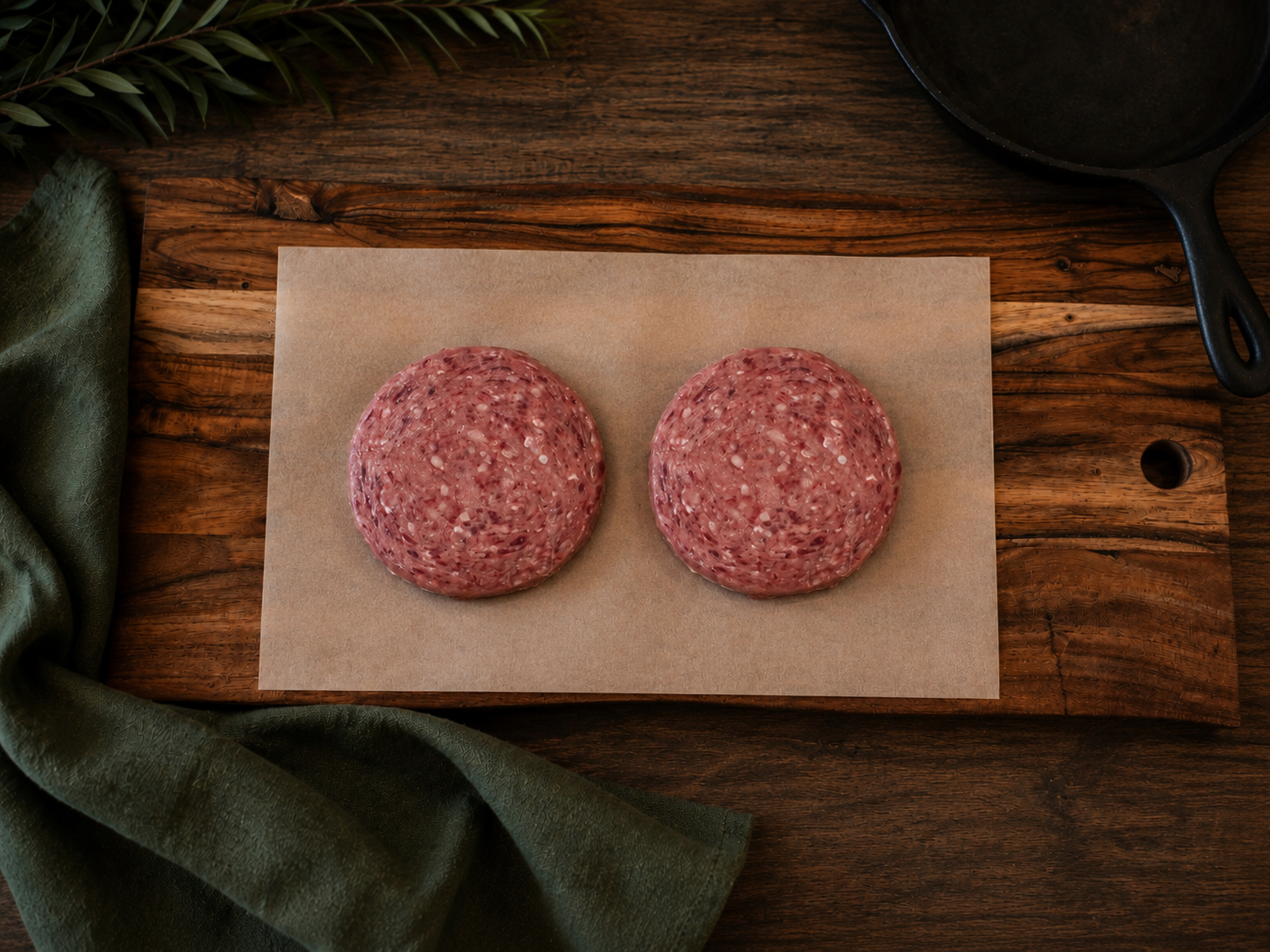 Two raw ground meat burger patties placed on a rectangular sheet of parchment paper, set on a rustic wooden cutting board with a dark green cloth and a black cast iron skillet in the background.